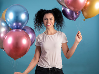 Portrait of young woman with black curly hair in casual dress smiling and holding party balloons isolated on blue background