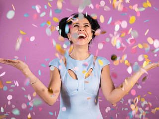Portrait of young woman with black curly hair in blue party outfit laughing and celebrating confetti rain isolated on pink background