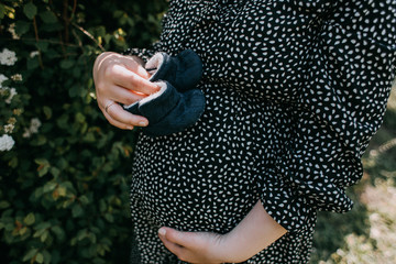 Belly of a pregnant girl on a background of greenery. The girl holds children's shoes in her hands. Waiting for the baby.