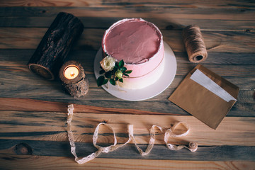 The cake is covered with white and pink cream, decorated with a white rose. Birthday cake. Cake stands on a wooden background with candles