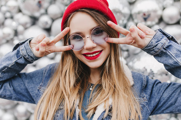 Close-up shot of enthusiastic blonde girl posing with peace sign in front of disco balls. Portrait of glad european woman in denim jacket expressing happiness.