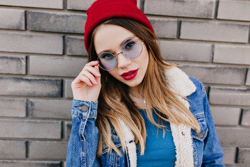 Close-up portrait of charming white girl touching her blue glasses and expressing interest. Outdoor photo of serious beautiful woman in denim jacket.