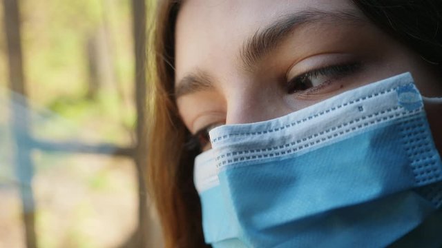 Close-up of a girl in a protective medical mask on her face sadly looks out the window while staying at home in quarantine