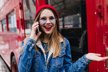 Surprised white girl in red hat talking on phone. Outdoor photo of romantic woman in denim jacket standing on the street with smartphone.