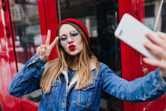 Lovely young woman with red lips making selfie in spring day and using phone. Outdoor portrait of ecstatic blonde girl in denim jacket taking pictures of herself with kissing face expression.