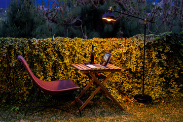 Night shooting a remote workplace in the form of a wooden chair and a table with a laptop and a glass and a bottle of red wine against a green hedge and illuminated by a floor lamp