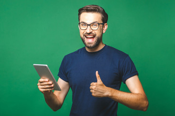 Winner! Happy young man in casual shirt and glasses standing and using tablet over green background. Thumbs up.