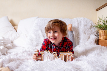 Obraz premium Happy little curly kid boy with smiley face lying on the bed at home. Stay at home quarantine coronavirus COVID-19 pandemic prevention. Wooden house inscription home.