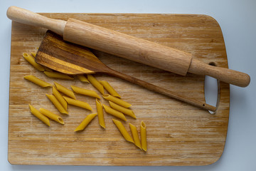 Top view of uncooked penne spaghetti with wooden rolling pin and wooden spatula on wooden cutting board