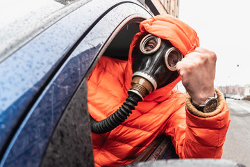 A man in a gas mask and an orange jacket with a hood sits behind the wheel of a blue car. Driver protected from coronavirus. The world in a pandemic.