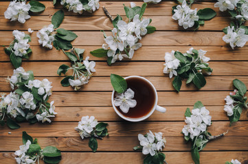 White cup of tea with petals of an apple tree on a wooden background, view from above.