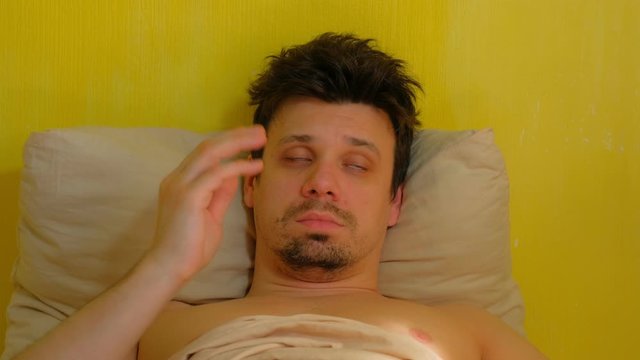 Man with hangover claps himself on cheeks to wake up lying in bed, portrait closeup. Tired young sleepy man in early morning. Yellow wallpaper background.