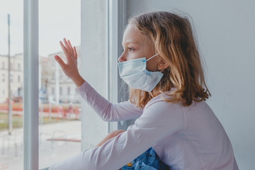 a sad European girl looks out the window with a coronovirus mask on her face. quarantine in Europe....