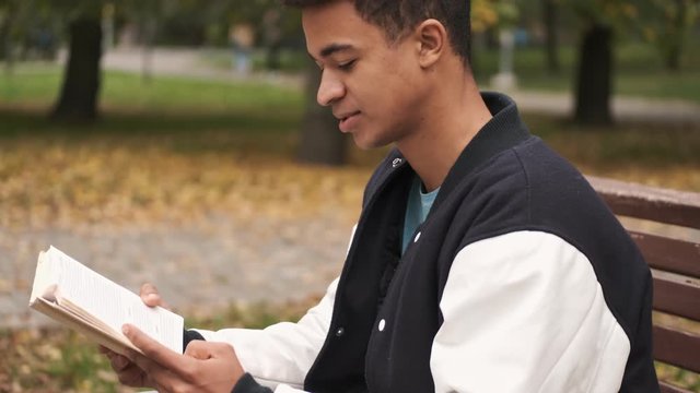 Concentrated young african guy student sit on bench reading book outdoors.