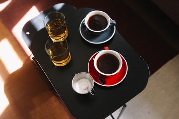 Top view on two cups of coffee, sugar and 2 glasses of whiskey on the black wooden table