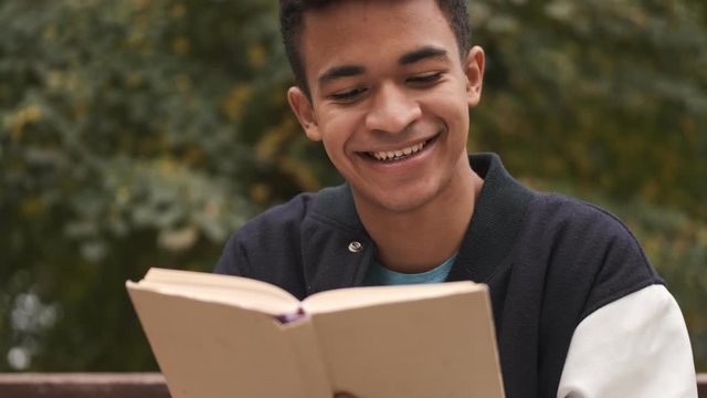 Concentrated young cheerful smiling african guy student sit on bench reading book outdoors.