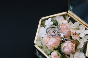 Vintage glass box with pink roses and white flowers and wedding rings lying on the black table background