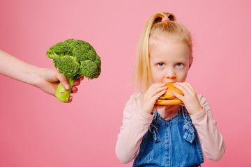 Hungry girl is choosing to eat a burger and dont want to choose broccoli is near to her on pink background. Unheathy vs healthy food. Concept of choice.