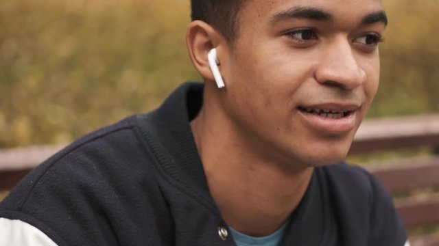 Handsome young positive african guy student listening music by wireless earphones outdoors in park.