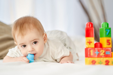 the baby is lying on bed with toys. He is gnawing a blue toy. Teething in a baby.