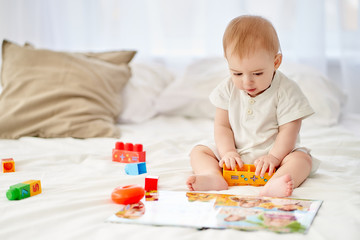 A little boy sitting on the bed plays with cubes. A book is lying beside him.