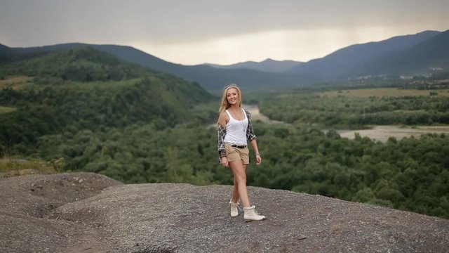 Happy cheerful stylish blonde girl, blogger, hipster in shirt and shorts walks singing, running, spinning, raising her hands to the side smiling. on the background of the landscape of the mountains