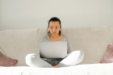 Asian woman dressed in grey shirt and trousers. Young Asian woman working from home while sitting on a sofa. Korean girl using laptop.