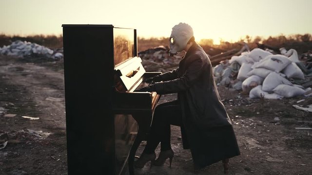 a girl in a black leather coat, in black gloves and a gas mask plays the piano among the ruins