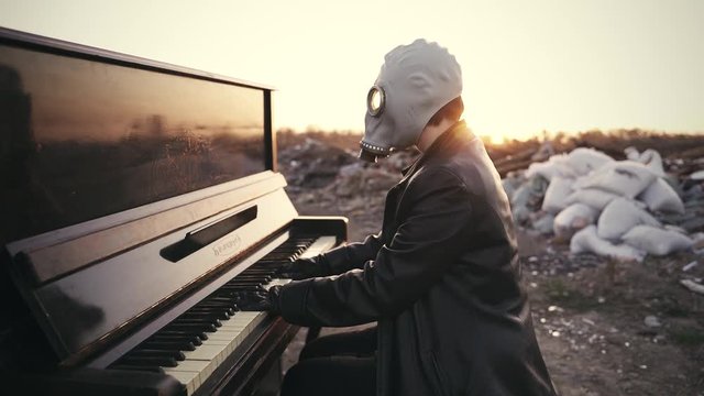 a girl in a black leather coat, in black gloves and a gas mask plays the piano among the ruins