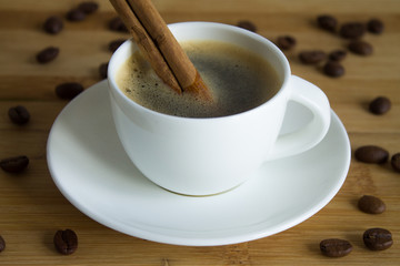 A small white espresso Cup on a saucer with a cinnamon stick on a wooden background with scattered coffee beans