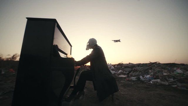 A girl in a black leather coat with black gloves and a gas mask plays the piano among the ruins. A plane is flying in the sky.