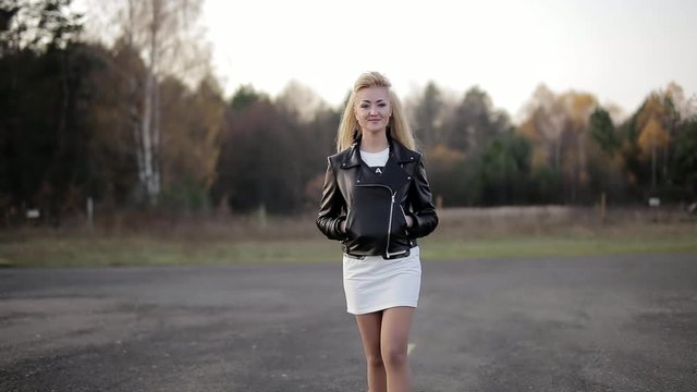 young blonde with long hair goes in black leather jacket and white short dress, at camera, smiling, hands in pockets, stylish attractive hipster, on nature background, and forest scenery