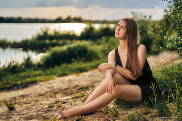 An attractive woman with bare feet and in a black dress sits on the sand and looks into the distance. Nature, water, green trees. The light of the sun falls on the surface of the water.