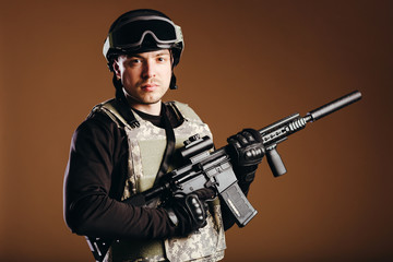 Soldier with gun is standing and looking on camera on brown background. Concept of war. Veterans, comrades, soldiers. Man in uniform.
