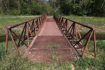 Rusty pedestrian bridge with grate floor as pathway across artificial water channel and triangular...