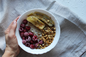 The girl's hand holds a plate of oatmeal porridge with raspberries and fried bananas. Healthy breakfast.