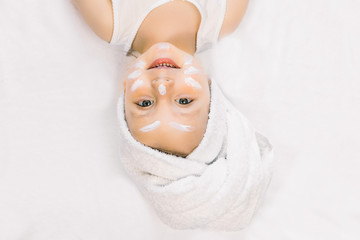 Cute little girl with hair wrapped in bath towel, lying on white bed with cream on her face after...