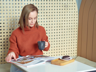 a girl in a brown jacket is in a cafe drinking coffee and reading a magazine.