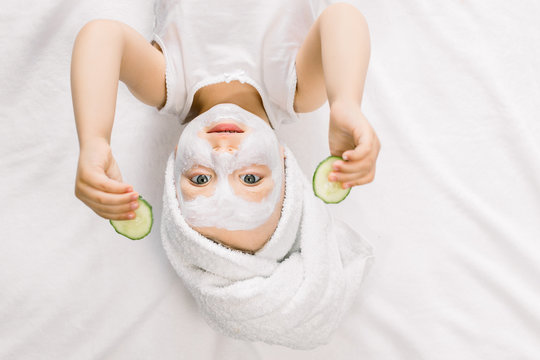 Funny little girl with a mask for skin face and towel on her head, holding two slices of fresh cucumber and looking at camera, while lying on white background after bath