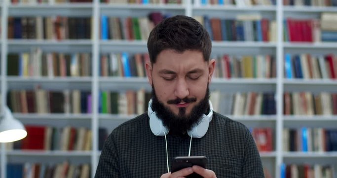 Young Cucasian male library worker or with headphone tapping and texting message on mobile phone. Man typing and scrolling on smartphone in bibliotheca. Book store costomer chatting with messages.