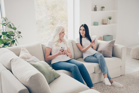 Portrait of two nice attractive lovely cheerful cheery women sitting on divan using digital device web app spending free time day weekend in white light interior house flat apartment
