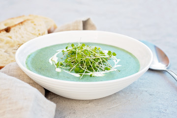 Cream soup of fresh spinach with cream and arugula microgreen in a white plate with ciabatta bread on a light gray background. Healthy and diet food.