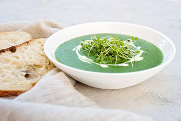 Cream soup of fresh spinach with cream and arugula microgreen in a white plate with ciabatta bread on a light gray background. Healthy and diet food.