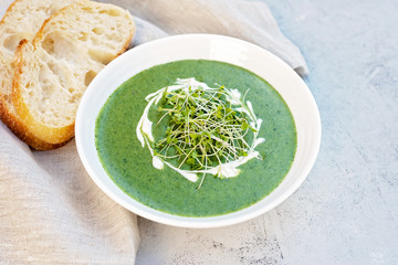Cream soup of fresh spinach with cream and arugula microgreen in a white plate with ciabatta bread on a light gray background. Healthy and diet food.