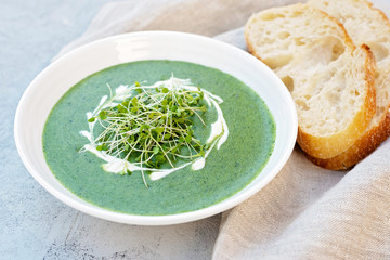 Cream soup of fresh spinach with cream and arugula microgreen in a white plate with ciabatta bread on a light gray background. Healthy and diet food.