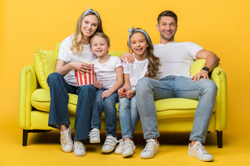 Obraz premium smiling parents and kids watching movie on sofa with popcorn bucket on yellow