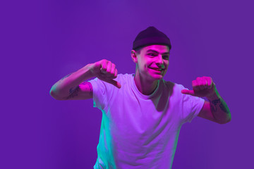 Naklejka premium Thumbs up, pointing. Caucasian man's portrait isolated on purple studio background in pink neon light. Beautiful male model in casual. Concept of human emotions, facial expression, sales, ad.