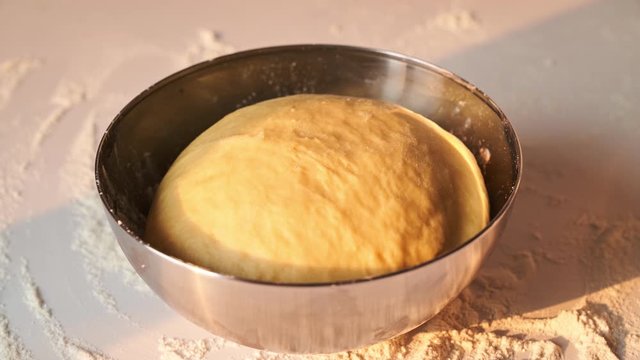 Rising fresh yeast dough in metal bowl on a table with sunlight and changing weather conditions. Horizontal timelapse video of dough fermenting and growing in a metal dish