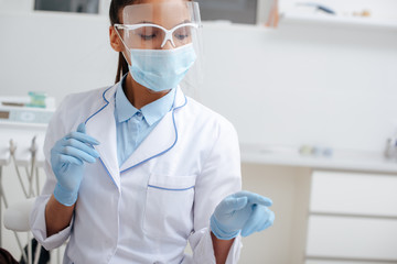 african american dentist in medical mask, face shield and latex gloves pointing with finger in clinic
