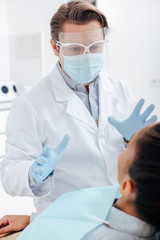 selective focus of dentist in face shield, medical mask and latex gloves gesturing near african american woman